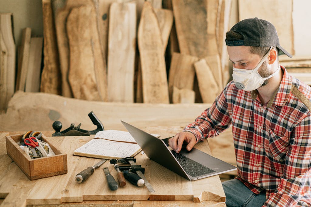 A craftsman wearing a mask, working on a laptop in a woodworking shop.