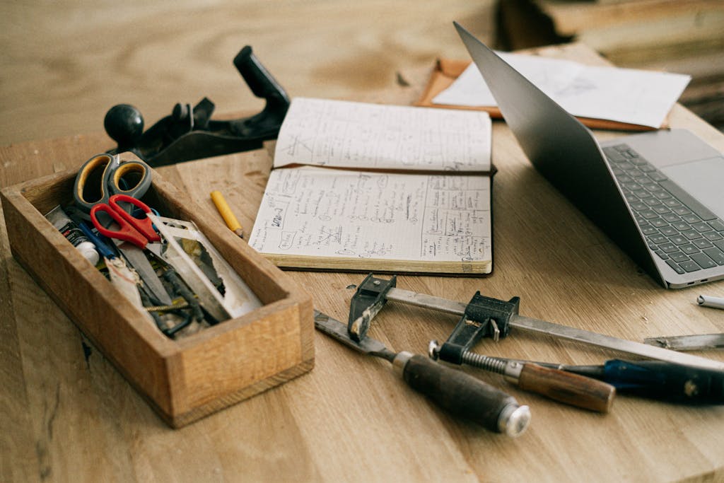 A creative woodworking workspace with tools, notebook, and laptop on a wooden table.