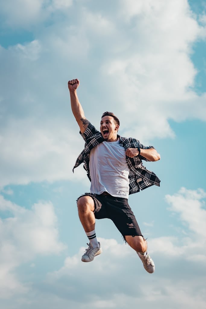 A joyful young man jumps midair with clouds and blue sky in the background, exuding energy and freedom.