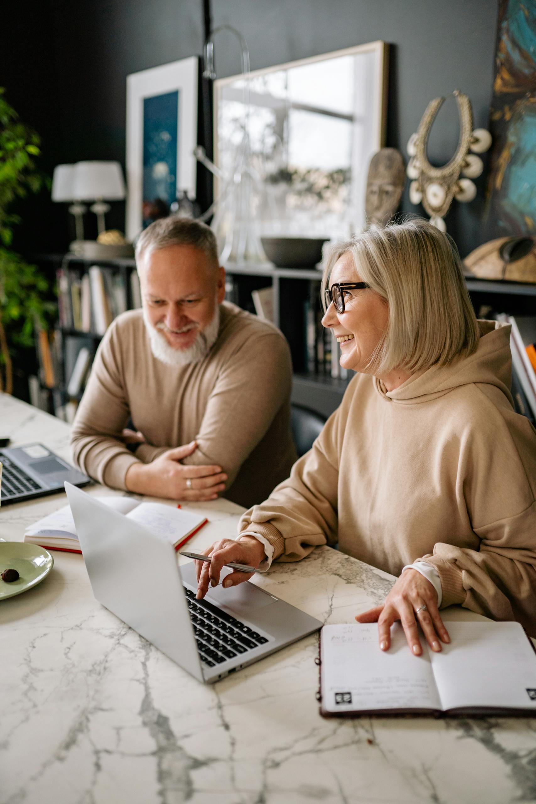 Senior couple using laptops at home, engaging in online activities with smiles and comfort.